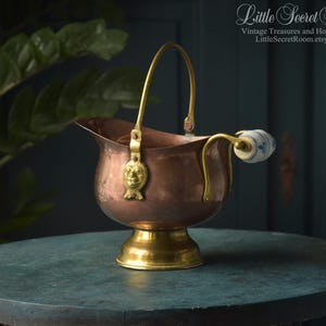 Puede incluir: Un cubo de carbón de cobre con base de pedestal de latón y asa de latón con un adorno de cabeza de león. El recipiente incluye un asa lateral con un agarre de cerámica blanca decorado con un patrón floral azul y el texto Little Secret Room.