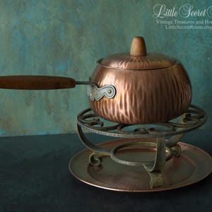 Può includere: Una pentola per fonduta in rame con manico e coperchio in legno, appoggiata su un supporto decorativo in metallo. La pentola ha una texture martellata ed è esposta su un piatto color rame abbinato. Lo sfondo è turchese e blu.