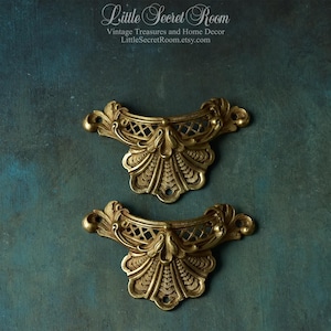 May include: Two matching brass-toned metal drawer pulls featuring an ornate, symmetrical design with a fan-shaped base, lattice detailing, and two mounting holes on each piece. The hardware has a textured, antique-style finish.