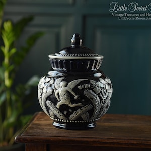 May include: A black ceramic jar with a lid, featuring a detailed relief design. The relief depicts a winged figure and foliage in white against the black background. The jar is displayed on a wooden surface.