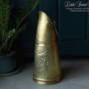May include: A vintage brass coal scuttle with a handle and ornate floral detailing. The scuttle has a conical shape and a curved top. The background includes a potted plant and a dark teal cabinet. The Etsy shop name "Little Secret Room" is visible.