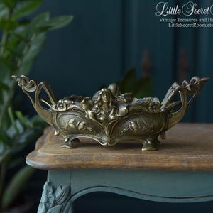 Puede incluir: Un cuenco decorativo antiguo de latón con una figura femenina central y motivos florales. El cuenco tiene asas ornamentadas y descansa sobre pequeños pies. El texto "Little Secret Room" es visible en la esquina superior derecha.