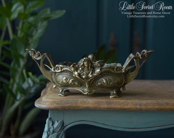 May include: An antique brass decorative bowl with a central female figure and floral motifs. The bowl has ornate handles and rests on small feet. The text "Little Secret Room" is visible in the upper right corner.