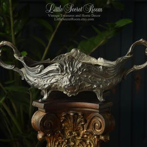 May include: An antique silver-toned decorative bowl with ornate handles and floral detailing. The bowl is displayed on a dark wooden pedestal with gold accents. The Etsy shop name "Little Secret Room" is visible in the image.
