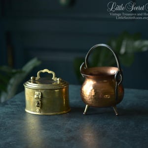 Könnte beinhalten: Eine ovale Messingdose mit Klappdeckel, zentralem Verschluss und kleinem Griff neben einem Kupferkessel mit gehämmertem Muster, drei kurzen Beinen und einem dunklen Metallhenkel. Der Text Little Secret Room ist deutlich zu sehen.