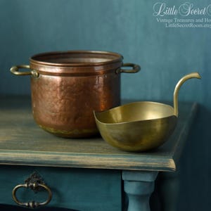 May include: A copper pot with two handles and a brass ladle with a curved handle sit on a teal surface. The pot has a hammered texture, and the ladle has a rounded bowl shape. The background is teal.