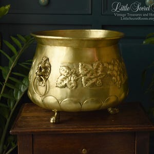 Può includere: Un'antica fioriera in ottone con maniglia ad anello a testa di leone e dettagli a vite. La fioriera poggia su tre piedini a zampa ed è esposta su un tavolo di legno. La fioriera è di colore dorato.