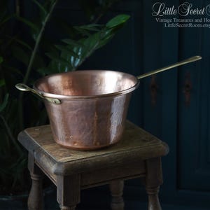 Op de afbeelding: Een vintage koperen kookpot met een lange messing handgreep en een kleine messing lus. De pot staat op een kleine houten kruk. De pot heeft een gehamerde textuur en een warme, roodbruine kleur.