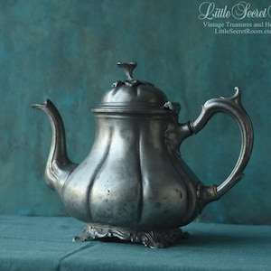 以下が含まれることがあります： 曲がった注ぎ口と装飾的なハンドルが付いた、アンティークシルバーのティーポット。 ティーポットはリブ付きのボディと小さなつまみ付きの蓋が付いています。 背景はティール色で、「Little Secret Room」の文字が見えます。