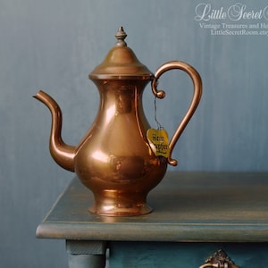 May include: A copper teapot with a shiny finish and a heart-shaped tag that says "Rein Apfer". The teapot is sitting on a blue and brown wooden table.