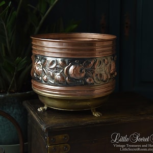 Op de afbeelding: Een koperen plantenbak met een decoratieve bloemenband en drie kleine voetjes. De plantenbak heeft een donkere patina en een gepolijste koperen rand. De plantenbak staat op een houten kist.