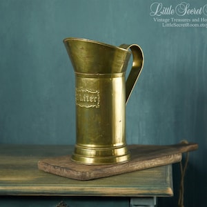 Può includere: Una brocca vintage in ottone con manico e beccuccio, appoggiata su un tagliere di legno. La brocca ha un corpo cilindrico e una parte superiore svasata. La parola "Liter" è impressa sul lato. Sfondo verde acqua.
