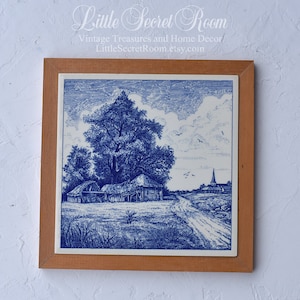 May include: A framed blue and white ceramic tile featuring a rural landscape scene. The artwork depicts a large tree, buildings, a road, and a church under a cloudy sky. The frame is made of light brown wood. Text at the top reads "Little Secret Room".