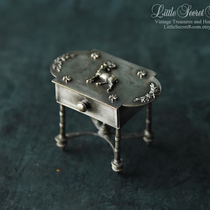 May include: A miniature silver-toned table with an oval top, adorned with floral and deer-shaped embellishments. The table features a small drawer with a round knob and ornate legs, showcasing a vintage aesthetic.