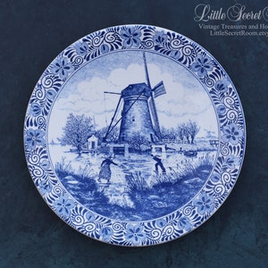 Może przedstawiać: Niebiesko-biały ceramiczny talerz z motywem holenderskiego wiatraka. Talerz przedstawia wiatrak, kanał i postacie w tradycyjnych holenderskich strojach. Brzeg talerza jest ozdobiony wzorem kwiatowym.