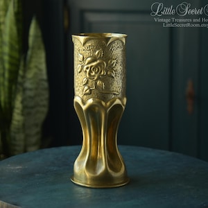 May include: A tall brass vase featuring a textured upper section with an embossed rose design and a smooth, fluted base. The cylindrical vessel has a scalloped rim and a metallic gold finish, displaying intricate floral detailing on its surface.