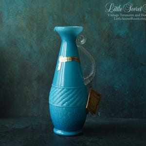 Op de afbeelding: Een turquoise glazen kan met een helder handvat en een decoratief touwontwerp in het midden. De kan heeft een gouden label en een hangend label. De achtergrond is donkerblauwgroen.