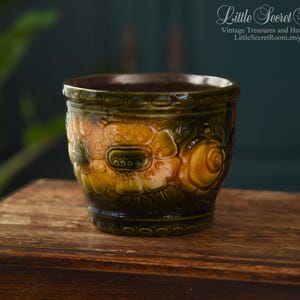 Może przedstawiać: Wazon ceramiczny w stylu vintage z kwiatowym wzorem w odcieniach zieleni, żółci i brązu. Wazon ma teksturowaną powierzchnię i stoi na drewnianym stole. Widoczna jest nazwa sklepu Etsy "Little Secret Room".