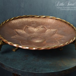 Könnte beinhalten: Ein Serviertablett aus Kupfer mit gehämmerter Oberfläche und einem dekorativen Blumendesign. Das Tablett hat einen geflochtenen goldenen Rand und drei goldene Tierfüße.