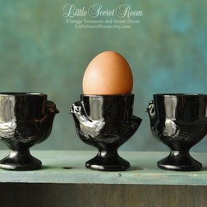 Pode incluir: Três porta-ovos pretos vintage com um design de pássaro, um deles segurando um ovo marrom. Os porta-ovos são feitos de um material brilhante e ficam em uma superfície verde clara desgastada. O fundo é de cor verde-azulado.