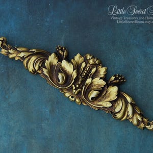 Puede incluir: Elemento decorativo dorado de estilo antiguo con diseño floral y de hojas. La pieza tiene una superficie texturizada y detallada, sobre un fondo verde azulado. El texto "Little Secret Room" es visible.