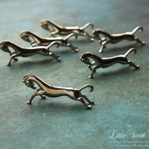 May include: Six small, silver-toned horse figurines are arranged on a textured, teal surface. The horses are in a running pose, with their legs extended. The figurines are detailed and reflective, catching the light.