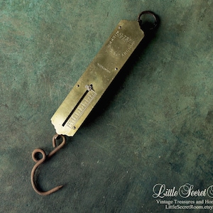 May include: A brass spring scale featuring a rectangular body with an integrated sliding indicator and a circular hanging loop at the top. A rusted iron S-hook is attached to the bottom. The metal surface displays engraved text including the word Warranted.