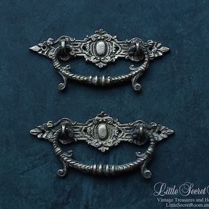 May include: Two antique silver drawer pulls with ornate designs. The pulls have a decorative scroll design and a central oval shape.