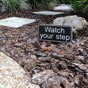 May include: A black and white sign with the text "Watch your step" on a brown mulch path.