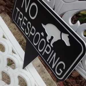 Op de afbeelding: Een zwart-wit bord met de tekst "NO TRESPOOPING" en een wit silhouet van een hond die poept. Het bord is bevestigd aan een wit metalen bankje.