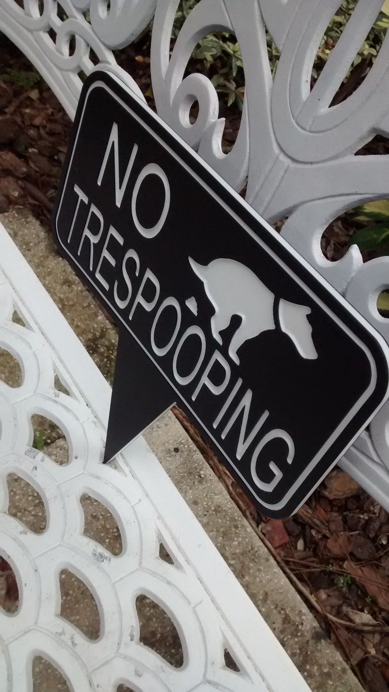 Op de afbeelding: Een zwart-wit bord met de tekst "NO TRESPOOPING" en een wit silhouet van een hond die poept. Het bord is bevestigd aan een wit metalen bankje.