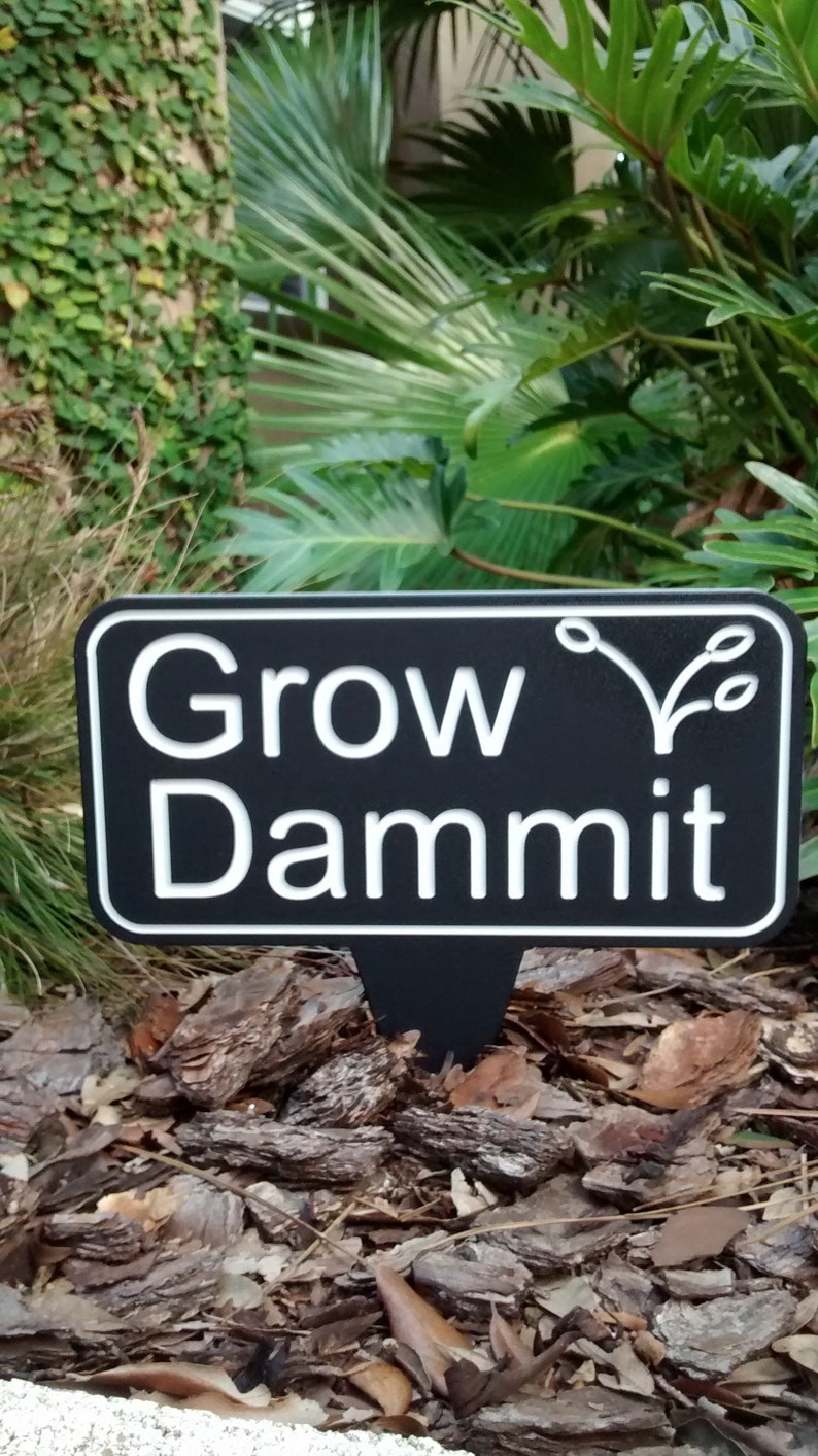May include: A black and white garden sign with the words "Grow Dammit" in a bold, sans-serif font. The sign features a decorative plant illustration and is set against a backdrop of mulch and greenery.