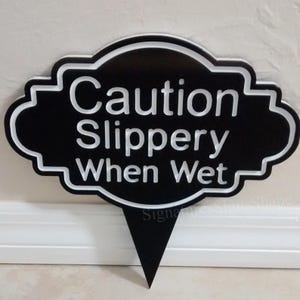 May include: Black and white sign with a white border and a pointed bottom. The sign reads "Caution Slippery When Wet".