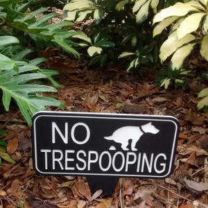 Op de afbeelding: Een zwart-wit bord met de tekst "NO TRESPOOPING" en een silhouet van een hond die poept. Het bord staat op een houten paal en bevindt zich in een tuin.
