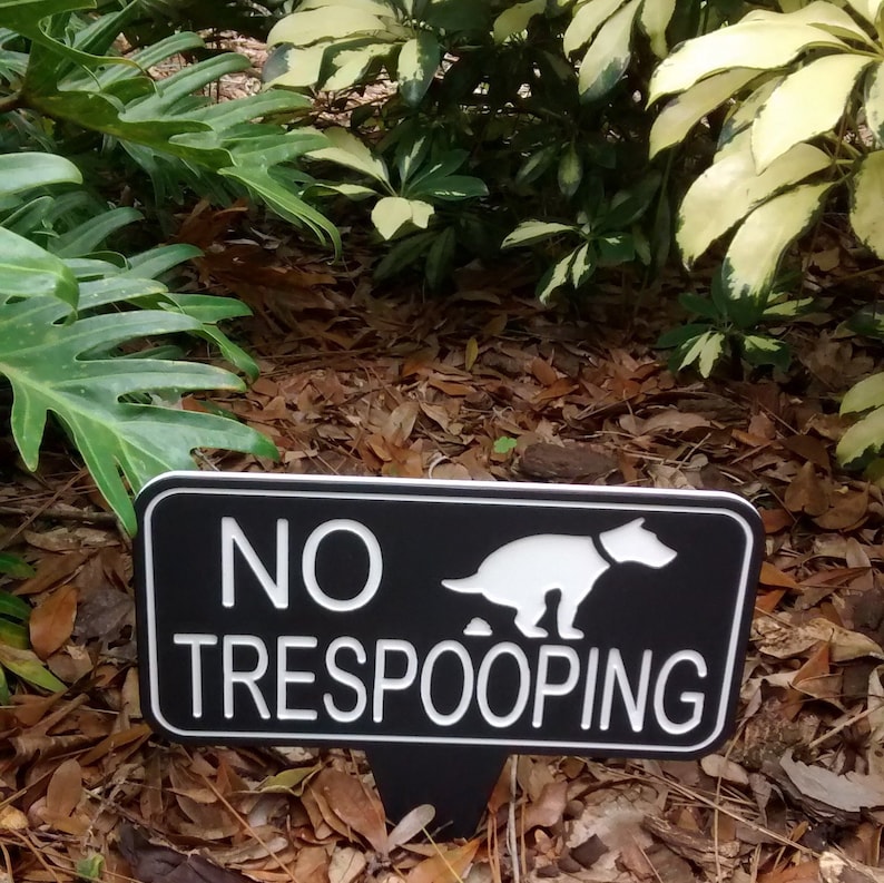 Op de afbeelding: Een zwart-wit bord met de tekst "NO TRESPOOPING" en een silhouet van een hond die poept. Het bord staat op een houten paal en bevindt zich in een tuin.