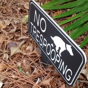 Op de afbeelding: Een zwart-wit bord met de tekst "NO TRESPOOPING" en een wit silhouet van een hond die poept. Het bord is gemaakt van zwart materiaal met een witte rand.