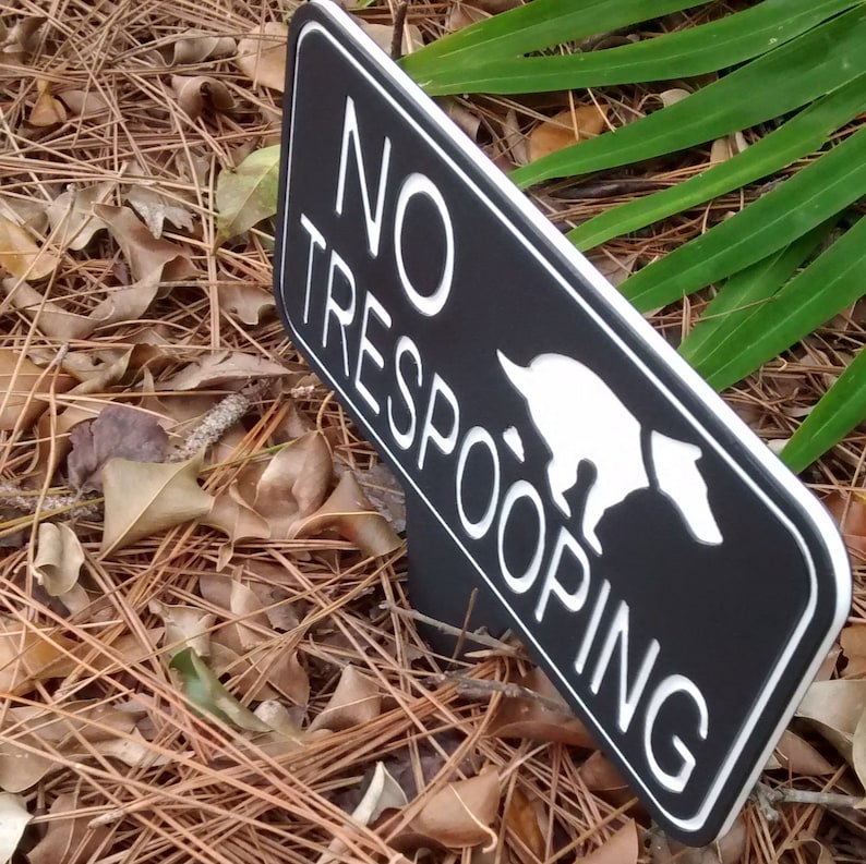 Op de afbeelding: Een zwart-wit bord met de tekst "NO TRESPOOPING" en een wit silhouet van een hond die poept. Het bord is gemaakt van zwart materiaal met een witte rand.