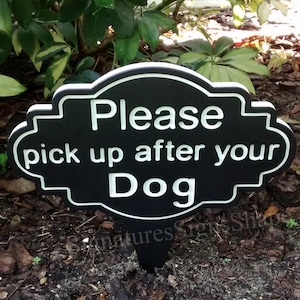 May include: Black and white sign with the text "Please pick up after your Dog" on a wooden stake. The sign is in a garden setting.