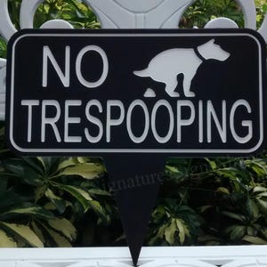 May include: Black and white sign with the text "NO TRESPOOPING" and a silhouette of a dog pooping. The sign is shaped like a pointer.