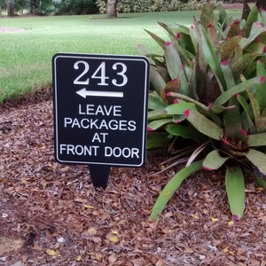 May include: A black sign with white lettering that reads "243 Leave Packages At Front Door" with an arrow pointing to the left.
