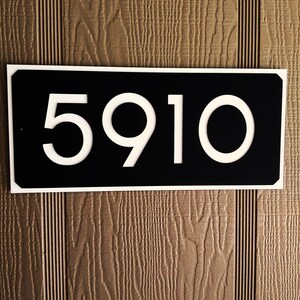 Pode incluir: Um sinal de n&uacute;mero de casa preto e branco com o n&uacute;mero 5910 em branco sobre um fundo preto. O sinal est&aacute; montado em uma superf&iacute;cie de madeira marrom.
