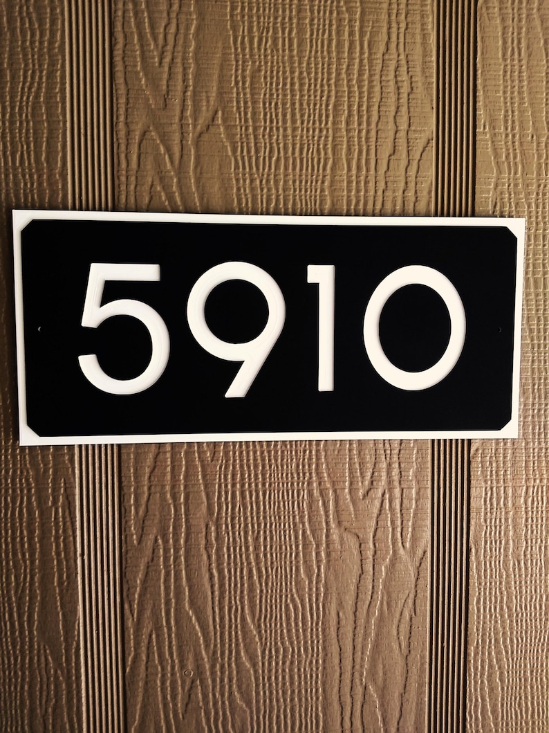 Pode incluir: Um sinal de n&uacute;mero de casa preto e branco com o n&uacute;mero 5910 em branco sobre um fundo preto. O sinal est&aacute; montado em uma superf&iacute;cie de madeira marrom.