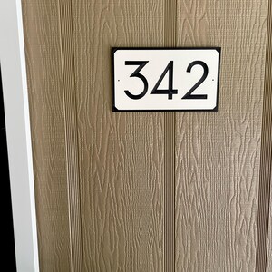Pode incluir: Um sinal de n&uacute;mero de casa retangular branco com borda preta e n&uacute;meros pretos. O sinal diz "342" e est&aacute; montado em uma parede de madeira marrom.