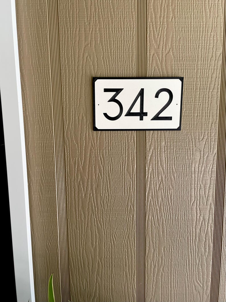 Pode incluir: Um sinal de n&uacute;mero de casa retangular branco com borda preta e n&uacute;meros pretos. O sinal diz "342" e est&aacute; montado em uma parede de madeira marrom.