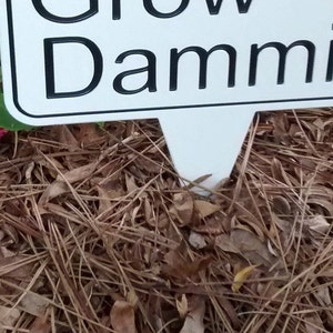 May include: A white sign with black text that reads "Grow Dammit" is placed in a garden bed. The sign is surrounded by brown mulch and dried leaves. Bright pink flowers and green leaves are visible in the upper left corner.