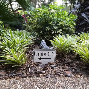 May include: A garden scene featuring a sign that reads "Units 1-3" with an arrow pointing left. A small, gray bird statue sits near the sign. The scene is surrounded by green and white plants and lush foliage.