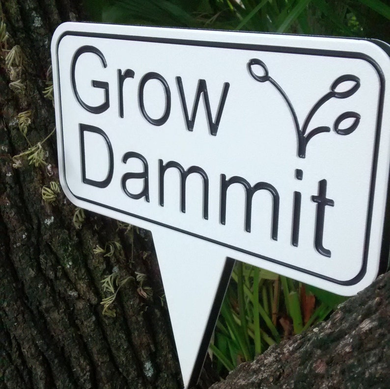 May include: A white garden sign with black text that reads "Grow Dammit." The sign has a black border and a stylized plant graphic. It is mounted on a tree trunk, with a triangular stake for ground placement.