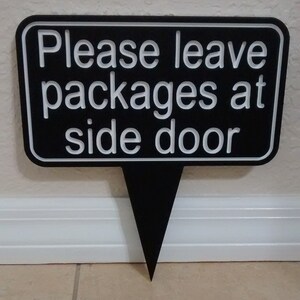 May include: Black sign with white text that reads "Please leave packages at side door". The sign is shaped like a speech bubble with a pointed bottom.
