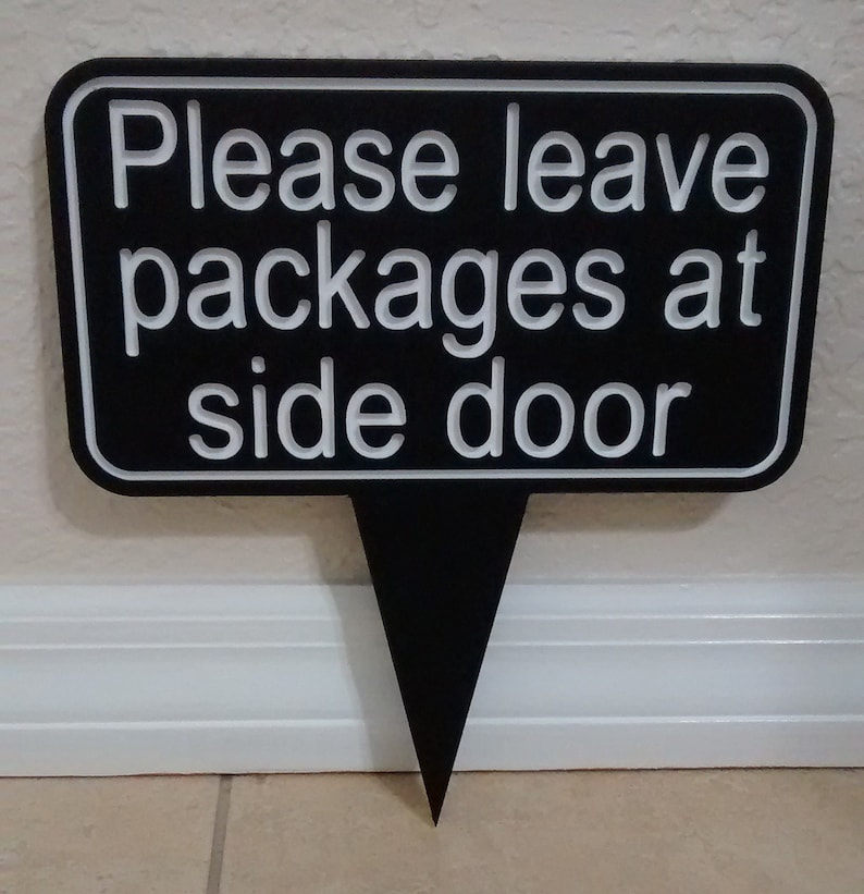 May include: Black sign with white text that reads "Please leave packages at side door". The sign is shaped like a speech bubble with a pointed bottom.