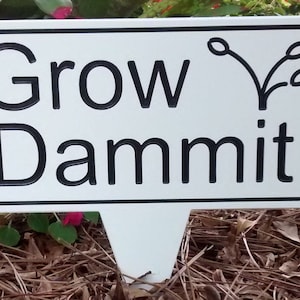 May include: A white garden stake with a black border and the words "Grow Dammit" in bold black letters. A stylized plant illustration is above the word "mit". The stake is in a garden setting with green foliage and pink flowers.